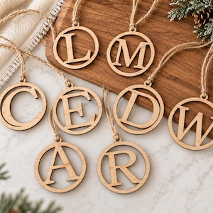 May include: A collection of eight circular wooden ornaments, each featuring a single cutout letter including L, M, C, E, D, W, A, and R. Each ornament is suspended by a thin, natural-colored jute twine loop, showcasing a light brown wood grain finish.