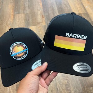 Puede incluir: Dos gorras de camionero negras con paneles traseros de malla. Una gorra presenta un logotipo con el texto "Barrier Island Marine" y un gr&aacute;fico de un barco y una gr&uacute;a. La otra gorra presenta el texto "Barrier" y un dise&ntilde;o de rayas horizontales en naranja, amarillo y rojo.