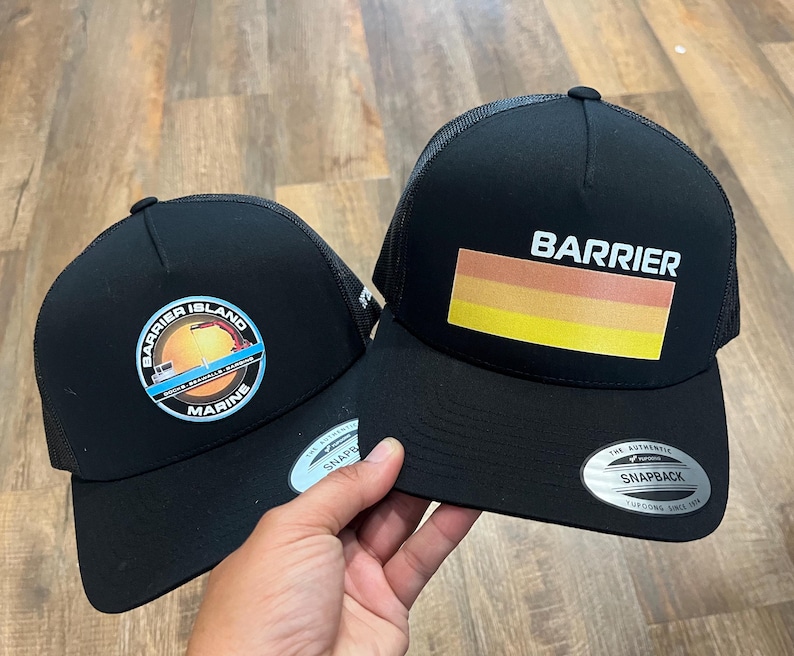 Puede incluir: Dos gorras de camionero negras con paneles traseros de malla. Una gorra presenta un logotipo con el texto "Barrier Island Marine" y un gr&aacute;fico de un barco y una gr&uacute;a. La otra gorra presenta el texto "Barrier" y un dise&ntilde;o de rayas horizontales en naranja, amarillo y rojo.