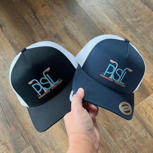 Puede incluir: Dos gorras de camionero, una negra y otra azul marino, con paneles de malla blanca. Ambas gorras tienen un logotipo blanco y turquesa que dice "PSL Paramount Streetlight".