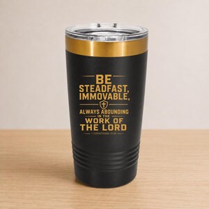 May include: Black tumbler with a gold rim and lid. The tumbler features the text "BE STEADFAST, IMMOVABLE, ALWAYS ABOUNDING IN THE WORK OF THE LORD" in gold lettering. The text is from 1 Corinthians 15:58.