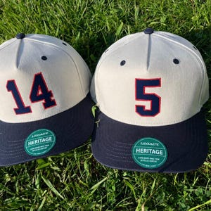 May include: Two baseball caps with off-white crowns and navy blue brims. The left cap features the navy blue number 14 with a red outline, and the right cap features the navy blue number 5 with a red outline. Both have green Legacy Heritage stickers.