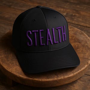 May include: A black baseball cap with the word "STEALTH" embroidered in purple across the front. The cap is made of a smooth material and has a curved brim. The hat is sitting on a wooden surface.