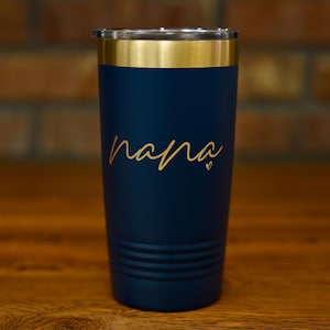 May include: Navy blue insulated tumbler with a gold rim and lid. The word "nana" is written in gold script on the front, with a small heart. The tumbler is cylindrical and sits on a wooden surface.