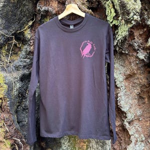 Peut inclure: Un t-shirt à manches longues gris foncé avec un motif rose d'oiseau dans un cercle et le texte "Badb Catha". Le t-shirt est suspendu à un cintre en bois. L'arrière-plan est une surface texturée.
