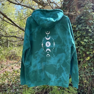 Peut inclure: Un sweat à capuche vert sarcelle avec un motif de phases lunaires blanches au dos. Le sweat à capuche a une capuche et un effet tie-dye délavé. Le motif des phases lunaires comprend des croissants de lune et une illustration centrale.