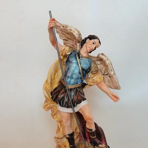 Könnte beinhalten: Eine Figur des Erzengels Michael, ein Kriegerengel mit Flügeln, der auf einem besiegten Dämon steht. Er ist mit einem Schwert und einem Schild dargestellt, und seine Flügel sind weit ausgebreitet. Die Figur ist aus bemaltem Harz und etwa 25 cm hoch.