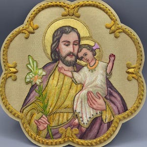 Puede incluir: Obra de arte religiosa bordada que representa a San José sosteniendo a un niño. La obra de arte tiene un halo dorado y un borde bordado dorado. Las figuras visten túnicas moradas, amarillas y blancas. El niño tiene un lazo rosa en el pelo.