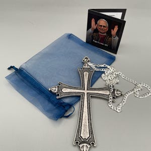 Könnte beinhalten: Ein silberfarbener Kreuzanhänger mit einem verzierten Design hängt an einer silbernen Kette. Ein blauer Kordelzugbeutel und eine kleine Karte mit einem Bild von Papst Leo XIV. sind ebenfalls vorhanden. Das Kreuz hat dekorative Details und eine strukturierte Oberfläche.