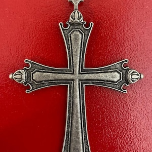 Op de afbeelding: Een zilverkleurige kruisvormige hanger met sierlijke details. Het kruis heeft een getextureerd oppervlak en decoratieve elementen aan de uiteinden van de armen en de bovenkant. Het hangt tegen een levendige rode achtergrond, wat het ontwerp benadrukt.
