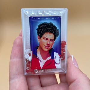 Peut inclure: Un boîtier rectangulaire en plastique transparent contenant un petit portrait d'une jeune personne aux cheveux bruns bouclés portant une chemise rouge et blanche. À l'intérieur, un chapelet de perles rouges et blanches entoure le portrait.
