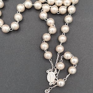 Könnte beinhalten: Ein Rosenkranz mit großen, runden, perlenartigen Perlen, die auf einer silbernen Kette aufgefädelt sind. Der Rosenkranz hat ein silbernes Kruzifix und einen kleinen silbernen Anhänger. Die Perlen sind cremeweiß.