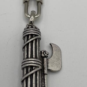 Può includere: Portachiavi color argento con un design stilizzato del fascio littorio, un fascio di verghe con una lama d'ascia. Il portachiavi ha un anello e un fermaglio per attaccarlo a chiavi o altri oggetti. Il design è dettagliato e metallico.