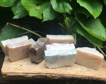 Peut inclure: Un assortiment de savons faits à la main de différentes couleurs, enveloppés dans du plastique transparent, présentés sur une planche de bois rustique. Les savons varient en nuances de marron, blanc et bleu, avec des textures visibles et certains estampillés de texte. Le fond est composé de feuillage vert.