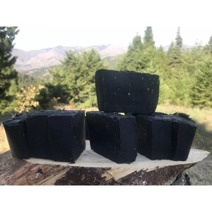 Peut inclure: Cinq savons noirs dispos&eacute;s sur une surface en bois. Le savon est dans un environnement naturel avec des arbres et des montagnes en arri&egrave;re-plan.