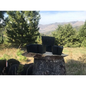 Peut inclure: Quatre barres de savon rectangulaires noires sont empil&eacute;es sur une souche d'arbre dans un cadre naturel. Le savon est devant une for&ecirc;t verte et une cha&icirc;ne de montagnes en arri&egrave;re-plan.