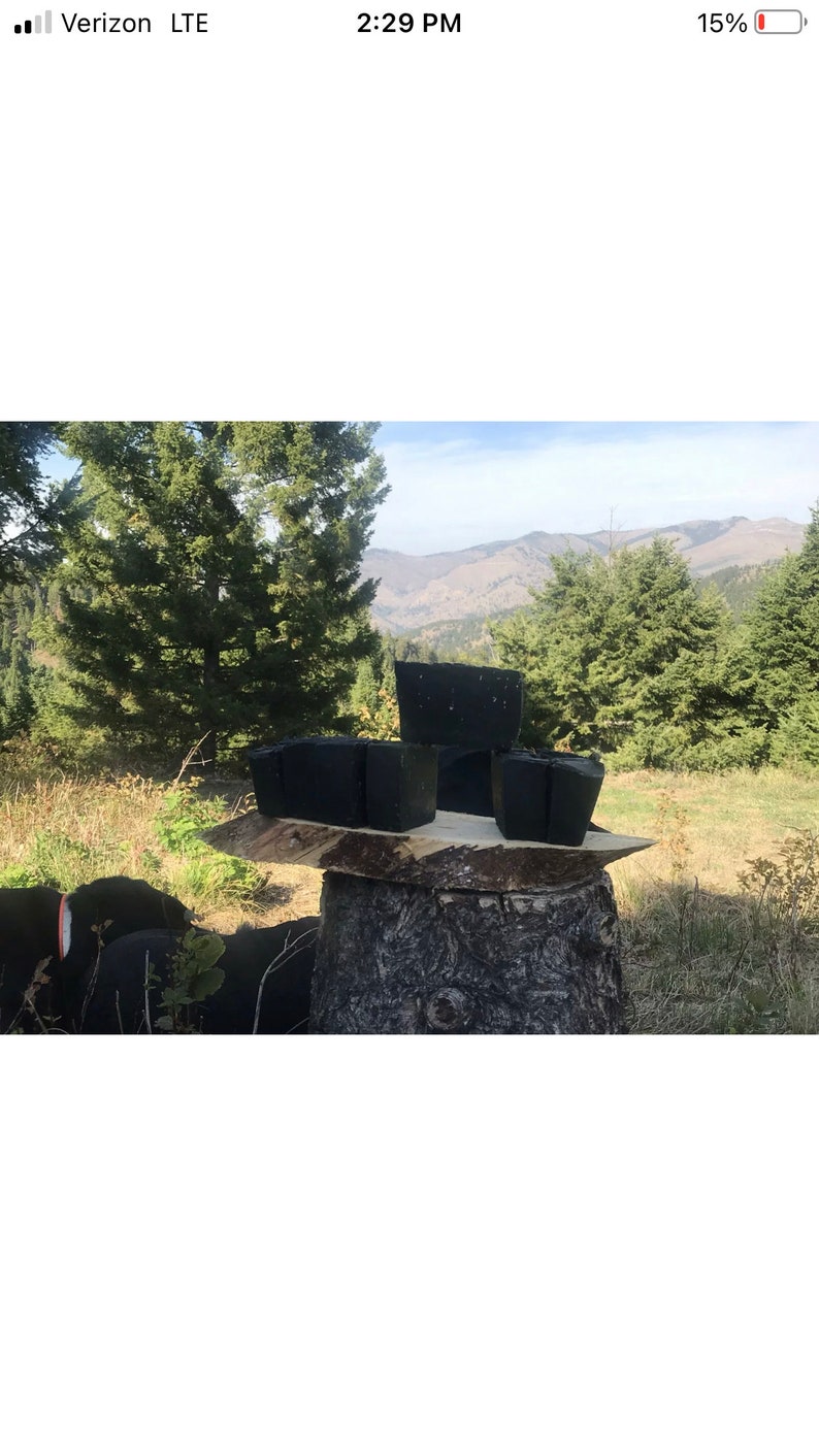 Peut inclure: Quatre barres de savon rectangulaires noires sont empil&eacute;es sur une souche d'arbre dans un cadre naturel. Le savon est devant une for&ecirc;t verte et une cha&icirc;ne de montagnes en arri&egrave;re-plan.