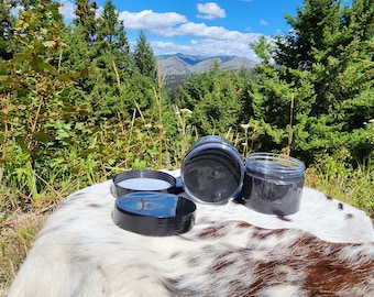 Peut inclure: Trois pots en plastique noir avec des couvercles, deux des couvercles sont enlevés, posés sur un tapis en peau de vache blanc et brun. Les pots sont nets et l'arrière-plan est une vue floue d'une forêt et de montagnes.