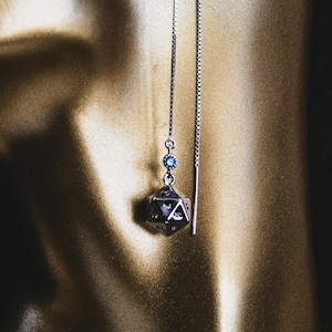 May include: A silver-toned threader earring featuring a dangling twenty-sided die charm with engraved star details and a small blue accent bead. The earring is suspended from a thin box chain that passes through the ear, creating a long, dangling effect.