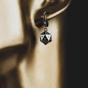 May include: A silver-toned hoop earring featuring a small, multi-faceted icosahedron charm. The charm is shaped like a twenty-sided die with clearly visible engraved numbers on its triangular faces. The earring is suspended from a metallic earlobe.
