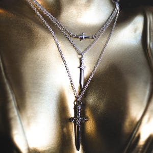 May include: A layered silver-toned necklace featuring three distinct chains of varying lengths. Each chain holds a small, detailed metal sword pendant. The pendants are arranged vertically, with the smallest sword at the top and the largest at the bottom.