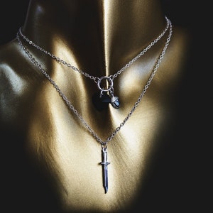 May include: A silver chain necklace with a black pendant and a silver sword pendant. The necklace is layered on a gold mannequin.