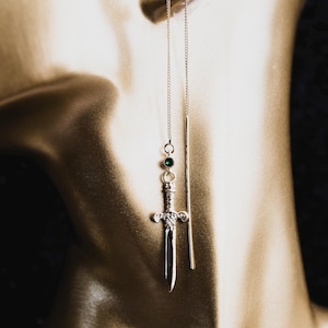 May include: A silver-toned threader earring featuring a delicate chain that suspends a small, round, dark green gemstone above a detailed miniature sword charm. The earring design includes a long, straight metal bar that hangs parallel to the chain.