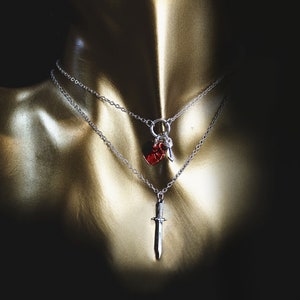 May include: Two silver necklaces with pendants. One pendant is a red heart shape and the other is a silver sword.