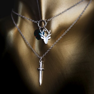 May include: Two silver necklaces with pendants. One pendant is a deer head and the other is a sword. The deer head pendant has a green crystal.