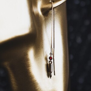 May include: Gold-toned bullet-shaped earring with a red gemstone accent. The earring hangs from a delicate chain, showcasing a unique and edgy design. The earring is shown on a mannequin.