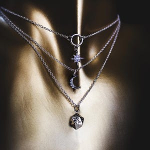 May include: A layered silver-toned necklace featuring three distinct chains. The top chain has a star charm, the middle chain displays a crescent moon pendant, and the bottom chain holds a multi-faceted geometric charm with engraved patterns.