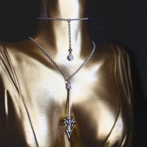 May include: A silver necklace with a long pendant shaped like a spear. The pendant has a detailed design and a small red gemstone at the top. The necklace is layered over a shorter silver choker with a small rose pendant.