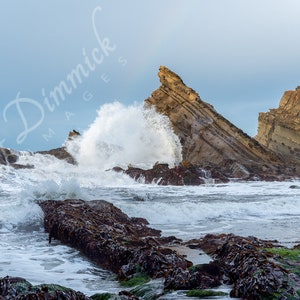May include: A powerful wave crashes against a rocky cliff face, creating a dramatic spray of water. The cliff is made of layered rock, and the water is a vibrant blue.