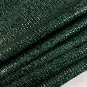 May include: A close-up of a dark green crocodile embossed leather. The leather is folded and textured, showing the intricate details of the embossed pattern.