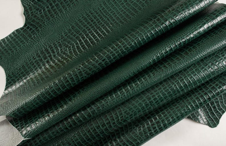 May include: A close-up of a dark green crocodile embossed leather. The leather is folded and textured, showing the intricate details of the embossed pattern.