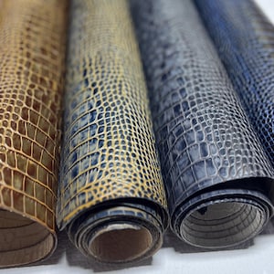May include: Rolled sheets of faux leather in shades of brown, blue, and gray. The material has a textured, crocodile-like pattern. The rolls are tightly wound, revealing the underside of the material.