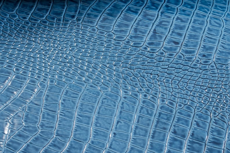 May include: Close-up of a blue crocodile-embossed leather texture. The leather has a subtle sheen and a repeating pattern of scales.