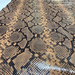 May include: Close-up of a brown and black snakeskin fabric with a glossy finish. The fabric has a repeating pattern of scales.