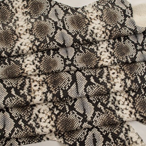 May include: Close-up of a fabric with a snakeskin pattern in shades of brown, black, and white. The fabric is folded and textured, creating a realistic snakeskin effect.