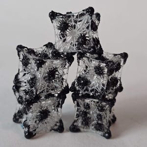 May include: A stack of three abstract, geometric sculptures. Each sculpture is a clear, cube-like form with a web-like structure and black, floral-like accents. The sculptures are arranged in a pyramid shape against a white background.