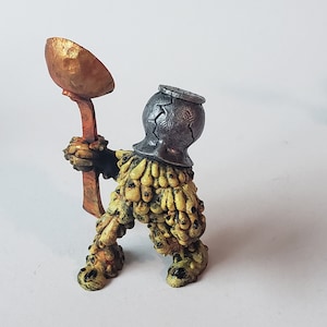 May include: A small, green, and yellow figurine with a pot on its head and a golden spoon in its hand. The figurine is covered in small, yellow bumps.