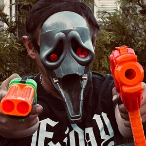May include: A person wearing a silver and black mask with red eye sockets, holding two orange and green toy guns. The person is wearing a black t-shirt with the word "Friday" in white script.