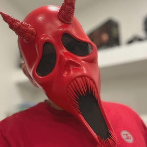 May include: A red devil mask with two textured horns, black mesh eye coverings, and a wide open mouth with sharp teeth. The mask is a solid red color and appears to be made of a hard material. The mask is worn by a person wearing a red shirt.