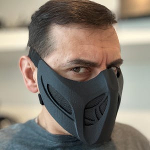 May include: A black face mask with a mesh design over the mouth and nose, secured by an adjustable strap. The mask has a futuristic, angular design, and is made of a textured material.