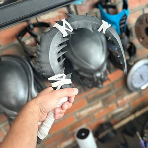 Może przedstawiać: Czarno-białe zakrzywione ostrze z ząbkowanym brzegiem i białą taśmą owiniętą wokół rękojeści. Ostrze jest trzymane w dłoni osoby.