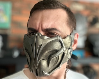 May include: A silver-coloured mask with a detailed, intricate design. The mask covers the nose and mouth and has a sharp, angular shape.