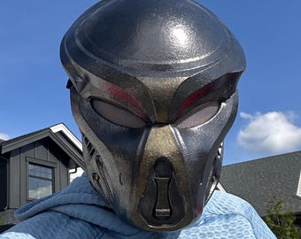 May include: A full-face helmet featuring a dark metallic finish with bronze-toned accents around the central nose and eye areas. The helmet has a smooth, rounded top, mesh-covered eye openings, and a vertical ventilation detail on the front.