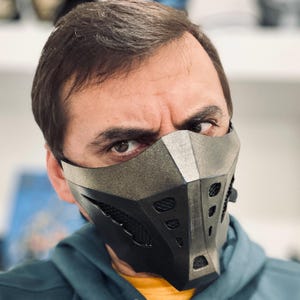 Op de afbeelding: Een zwart en grijs gezichtsmasker met een geometrisch ontwerp. Het masker heeft een getextureerde, metallic uitstraling en beschikt over ventilatiegaten. Het masker bedekt de onderste helft van het gezicht en is vastgezet met bandjes.