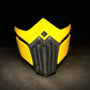 Può includere: Una maschera facciale gialla e nera con un design geometrico. La maschera ha un motivo nero a griglia sulla zona della bocca e del naso.