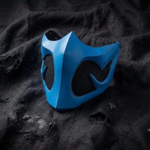 May include: A bright blue face mask featuring a rigid, angular design with a pointed chin and two large, symmetrical cutouts. The cutouts are filled with a black, fine-mesh fabric, providing a contrast to the smooth, solid blue surface of the mask.